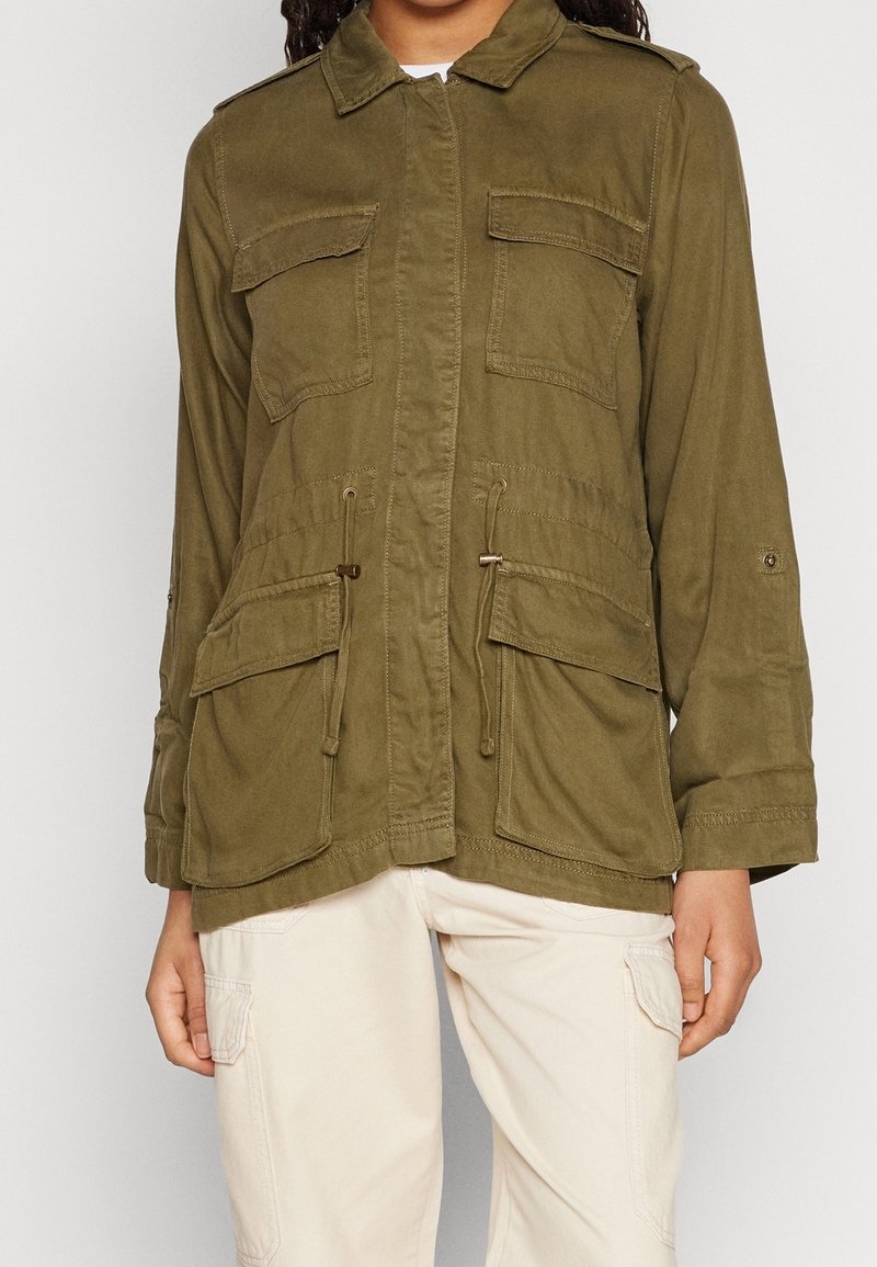 Oliwkowo-zielona kurtka użytkowa z przednimi kieszeniami na klapę, talią ze ściągaczem oraz beżowe spodnie cargo z bocznymi kieszeniami.