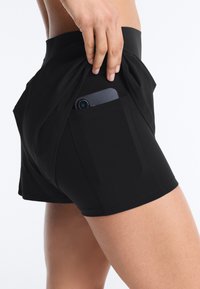 Kéz egy okostelefont helyez a fekete sportnadrág oldalzsebébe, amelyet egy személy a felső combján és csípőjén visel.