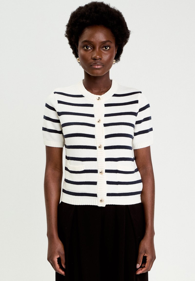 Mujer con cabello corto y rizado que lleva un cárdigan blanco de manga corta con botones y rayas horizontales negras, junto con una falda negra, sobre un fondo liso.