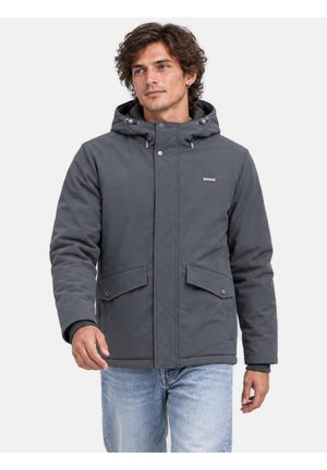 Homme portant une veste d'hiver à capuche gris foncé avec des poches à rabats et un jean bleu clair, debout devant un fond clair uni.