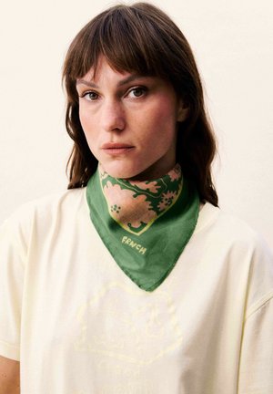 Femme aux cheveux bruns portant un bandana vert à motifs floraux autour du cou et une chemise de couleur claire sur un fond uni.