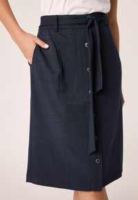 La gonna blu navy presenta un nastro in vita, tasche laterali e dettagli a bottoni lungo la parte anteriore. Il materiale ha una consistenza liscia con una leggera lucentezza.