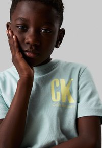 Camiseta de algodón azul claro con un gran y audaz gráfico en dorado de "CK" y el texto "Calvin Klein Jeans". Textura suave; mangas cortas.