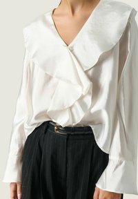 Blusa de satén blanca con un escote en V profundo y detalles de volantes en capas. Mangas largas con puños de volantes, combinada con pantalones negros de rayas diplomáticas a medida.