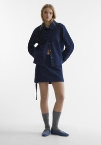 Giacca a fantasia navy con dettaglio floreale, mini gonna coordinata, calze alta fino al ginocchio grigie e scarpe blu slip-on. Texture del tessuto liscia.