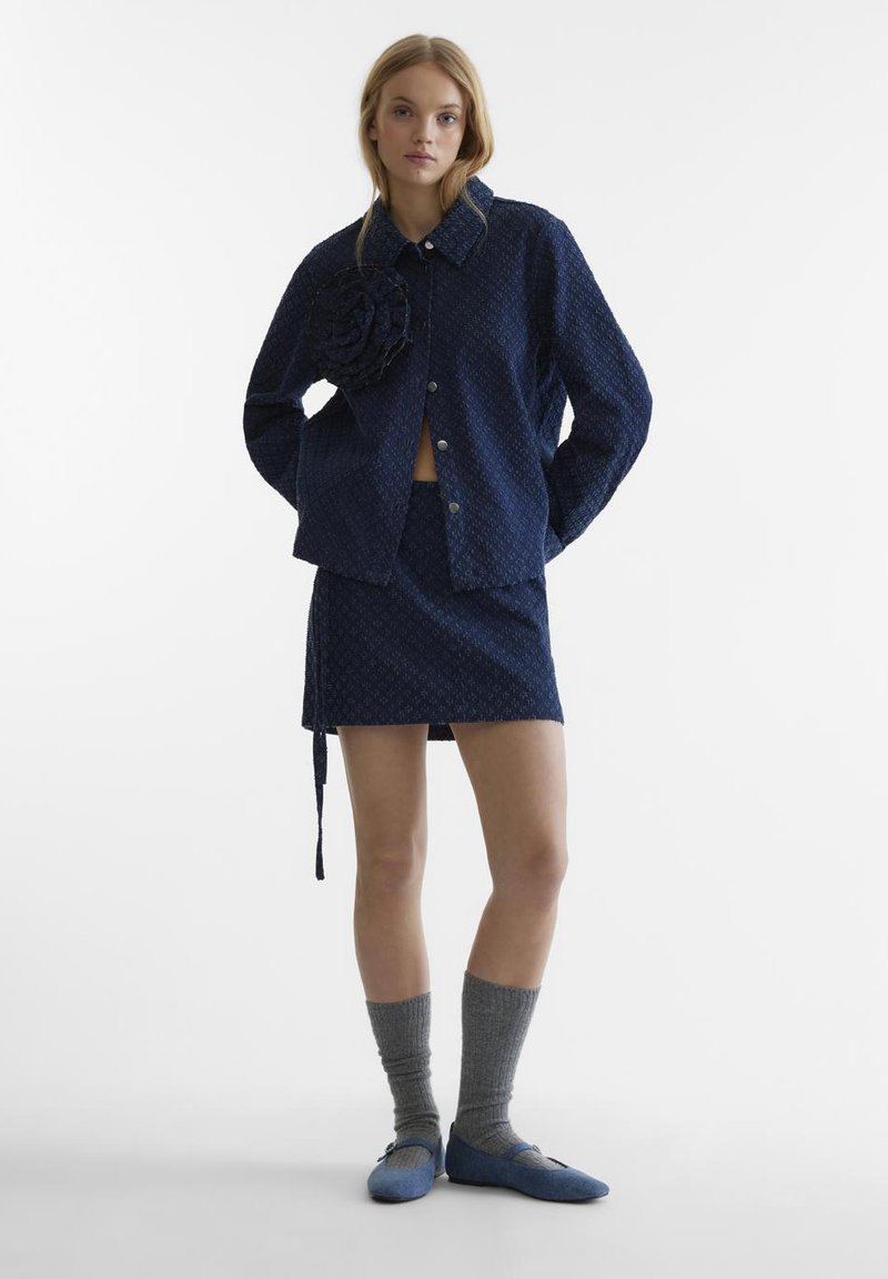 Giacca a fantasia navy con dettaglio floreale, mini gonna coordinata, calze alta fino al ginocchio grigie e scarpe blu slip-on. Texture del tessuto liscia.