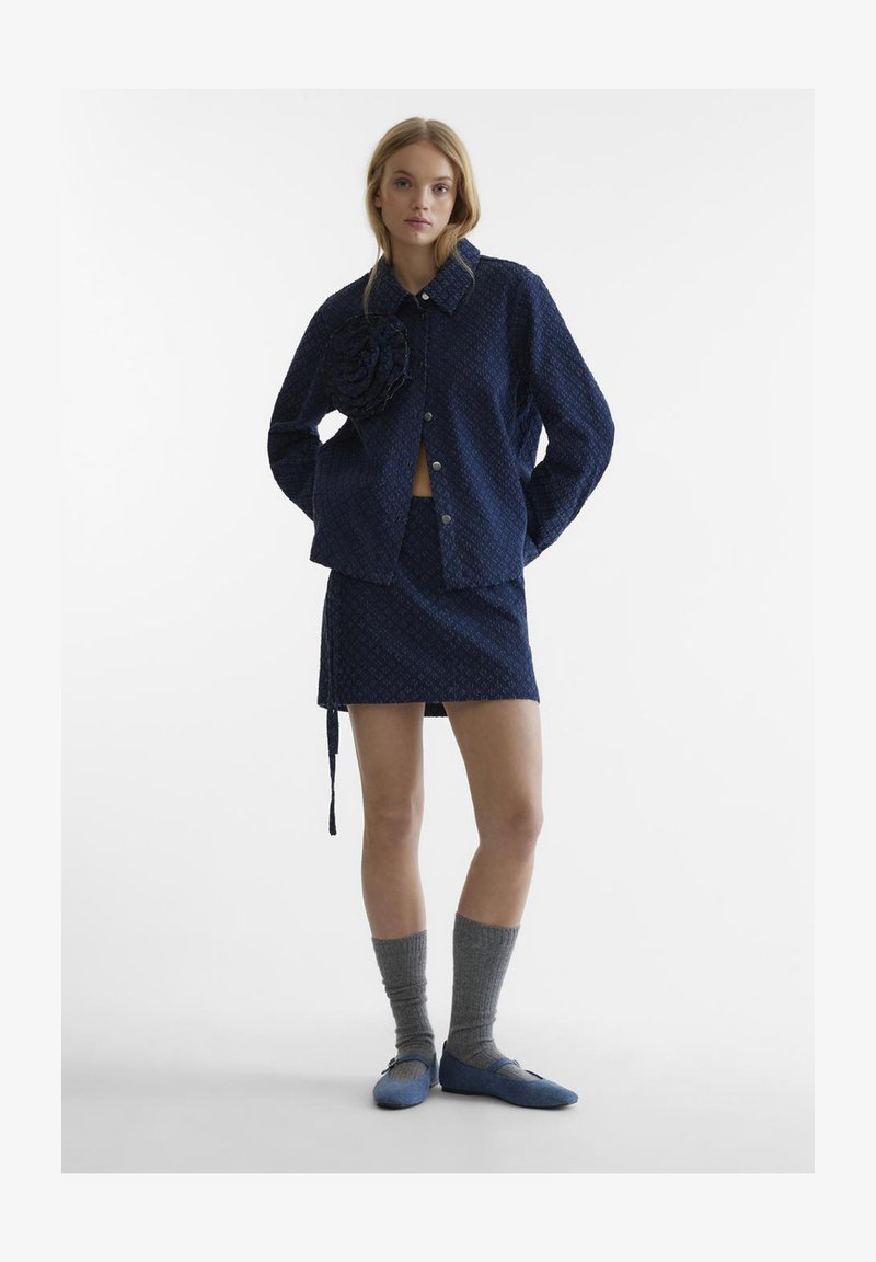 Giacca a fantasia navy con dettaglio floreale, mini gonna coordinata, calze alta fino al ginocchio grigie e scarpe blu slip-on. Texture del tessuto liscia.