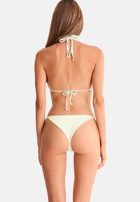 Femme aux longs cheveux portant un bikini blanc texturé avec un col halter et noué dans le dos, debout, dos à nous, sur un fond uni.