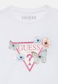 Άσπρη βαμβακερή μπλούζα με τριγωνικό γκλίτερ σχέδιο με λουλούδια και πεταλούδες σε παστέλ χρώματα, και το κείμενο «GUESS U.S.A.» σε ροζ.