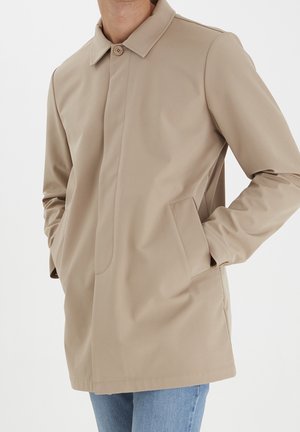 Uomo che indossa un cappotto beige monopetto con colletto e tasche, abbinato a jeans azzurri, con le mani nelle tasche del cappotto.