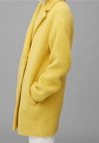 Abrigo de lana amarillo con una textura suave y esponjosa, que cuenta con un collar con muesca, dos bolsillos frontales y un corte relajado.