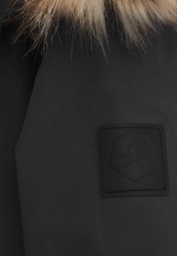 Svart tyg med matt yta, med en texturerad lapp med en logotyp. Lappen har en upphöjd emblem med en sköldliknande form.