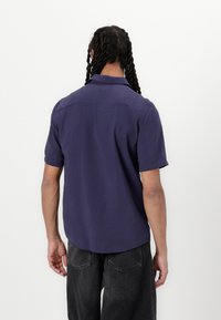 Kortærmet, skjortekrave skjorte i dyb marineblå stof, med lige kant og minimal detaljer bagpå. Let vævet tekstur.