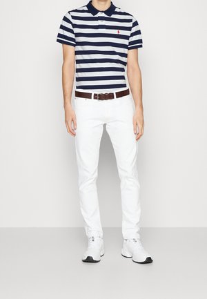 Mand iført navyblå og hvide vandrette stribede polo-shirt, hvide bukser, brunt bælte og hvide sneakers, stående mod en ensfarvet baggrund.