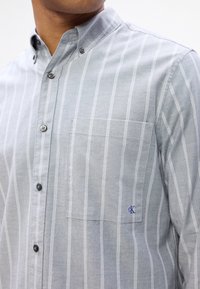 Pelēka svītrainā krekla ar apkakli; satur vienu krūšu kabatu un mazu zilu logotipu; izgatavots no teksturēta auduma.