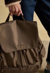 Main tenant un sac à dos marron Joules par la poignée supérieure, personne portant un jean foncé et une veste avec un poignet en polaire.