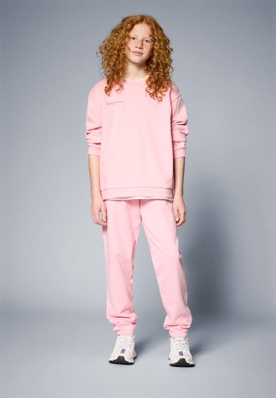 Rosa sweatshirt og matchende joggebukser, begge laget av mykt stoff. Har rund hals og løs passform. Brukt med hvite treningssko.