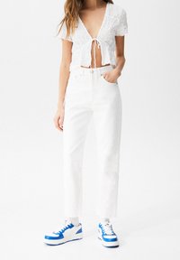 Crop top blanc texturé avec un nœud à l'avant, associé à un jean blanc taille haute à jambes droites et des baskets blanc-bleu.