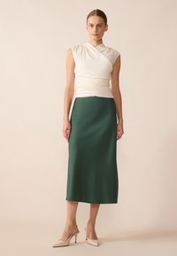 Top en satin blanc drapé avec des détails froncés, associé à une jupe longue en satin vert. La mannequin porte des talons ouverts beiges.