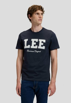 Junger Mann mit lockigem Haar, der ein schwarzes T-Shirt mit großem weißen "LEE" und "American Original" Text trägt, vor einem schlichten grauen Hintergrund stehend.