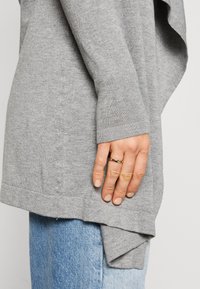 Light gray sweater with a relaxed fit, ribbed cuffs, and a slightly longer back hem. Worn over blue denim. Includes a ring on the hand.