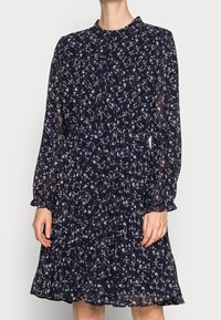 Navy sininen kukallinen mekko pitkillä hihoilla, laskostetulla hameella ja korkealla kaula-aukolla. Valmistettu kevyestä, läpikuultavasta kankaasta, jossa on hienovarainen kuvio.