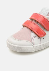 Zapatilla blanca con acentos de ante rosa y cuero sintético, que presenta dos correas de Velcro rojas. Suela de goma texturizada con patrón en el dibujo.