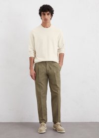Kremowy sweter z dzianiny, oliwkowe spodnie, beżowe buty. Model stoi prosto z rękami w kieszeniach na jednolitym tle. Minimalistyczny design.