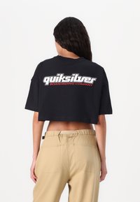 T-shirt nera cropped con logo "quiksilver" in bianco e rosso, maniche corte e vestibilità rilassata. Abbinata a pantaloni beige.