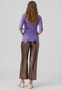 Fialová čipkovaná blúzka s tromi štvrtinovými rukávmi, kombinovaná s lesklými bronzovými širokými nohavicami. Modelka má na sebe svetlé sandále na blokovom podpätku.