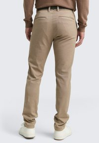 Homme portant un pantalon beige slim-fit, un pull marron à manches longues et des baskets blanches, se tenant de dos contre un fond clair.