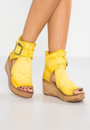 Ben, der bærer gule læder open-toe wedge sandaler med store side spænder, står på et lyst gulv mod en hvid baggrund.