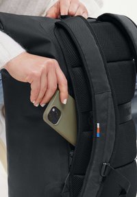 Svart ryggsäck med meshdetaljer, sidoficka som håller ett olivgrönt mobilfodral, med en flerfärgad logotyp på remmen.