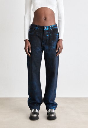 Ciemne, przylegające dżinsy wąskiego kroju z niebieskimi metalicznymi plamkami, noszone z czarno-białymi trampkami i białym, dopasowanym, prążkowanym sweterkiem ze skróconym krojem.