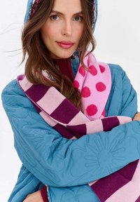 Prošívaná modrá bunda so vzorom kvetov, preložená cez bordový sveter, zahalená ružovo-fialovou šatkou s pruhmi a bodkami.