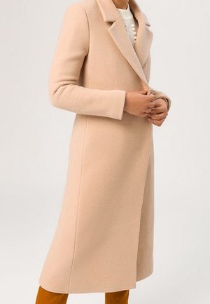 Femme portant un long manteau en laine beige par-dessus un pull blanc à gros boutons, se tenant devant un fond uni.