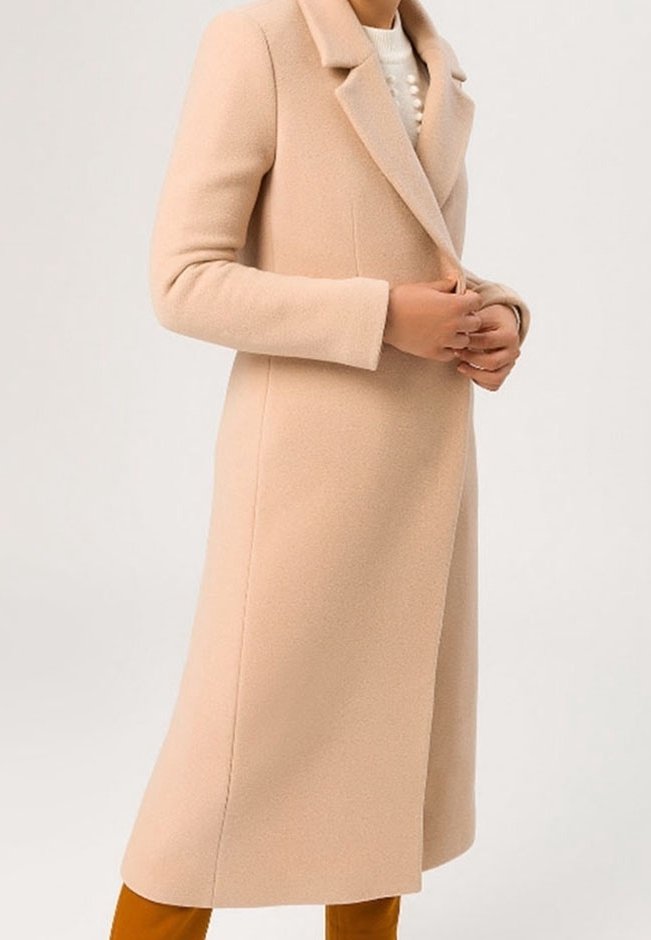 Femme portant un long manteau en laine beige par-dessus un pull blanc à gros boutons, se tenant devant un fond uni.
