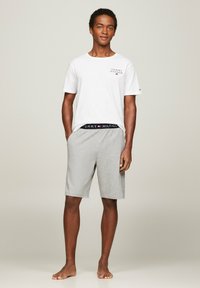 Witte kortarmige t-shirt met een klein logo, gecombineerd met lichtgrijze shorts met een zwarte merkband. Beroepen op blote voeten.