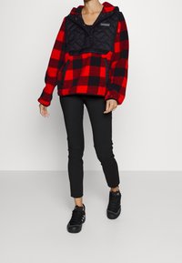 Czerwona i czarna kraciasta bluza z polaru z czarną pikowaną górną częścią i kapturem, połączona z czarnymi dopasowanymi spodniami i czarnymi trampkami.