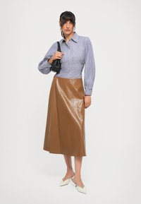 Camisa de botones a rayas azul y blanca con mangas abullonadas, combinada con una falda midi brillante marrón y mules de tacón blanco. Acento de bolso negro.