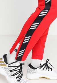 Pantalons de sport rouges avec un panneau latéral à rayures noires et blanches et des zips aux chevilles, portés avec des chaussettes blanches Adidas et des chaussures de sport blanches.
