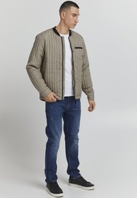 Giacca bomber trapuntata beige con colletto nero, tasca con zip frontale, indossata sopra una camicia bianca e abbinata a jeans blu strappati e sneakers nere.