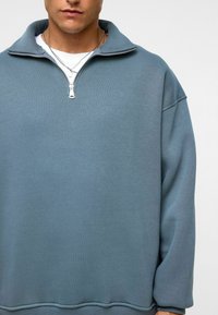 Lichtblauwe fleece sweatshirt met een hoge kraag en een halve ritssluiting. Heeft verlaagde schouders en een ontspannen pasvorm. Zachte textuur.