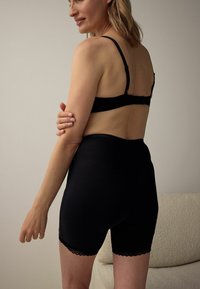 Fekete melltartó és magas derekú testformáló rövidnadrágok, szegélyükön csipkedíszítéssel és sima, rugalmas anyagból. A modell hátulnézetben látható.