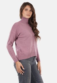 Maglione a collo alto di colore viola chiaro in morbido tessuto a maglia, con orli e polsini a costine. Il modello lo indossa con jeans scuri, posando di lato.