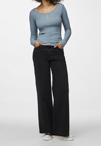 Kvinna klädd i en blå långärmad ribbad topp, vida svarta byxor och vita plattformsskor, står med händerna i fickorna.