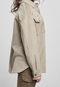 Boční pohled na osobu v béžové oversized košili s dlouhým rukávem, knoflíky a kapsou na prsou, která má na sobě hnědé cargo kalhoty.