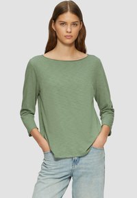 Grünes Langarmshirt mit Bootsausschnitt und lässiger Passform. Hat eine glatte Textur und abgerundeten Saum, kombiniert mit hellblauen Jeans.
