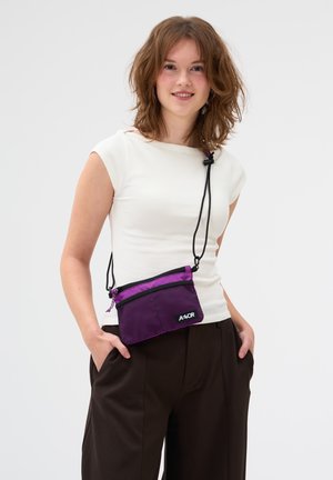 Junge Frau mit schulterlangem Haar, die ein weißes Oberteil, dunkle Hosen und eine kleine lilafarbene Umhängetasche trägt, mit den Händen in den Taschen, lächelnd.