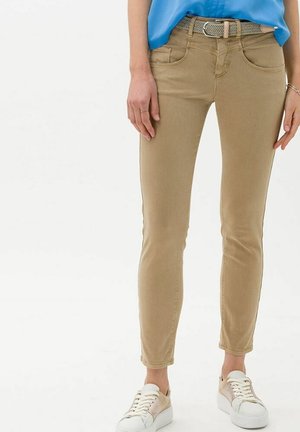 Woman wearing slim-fit beige pants, woven belt, blue shirt, and white sneakers, standing with arms relaxed by sides.